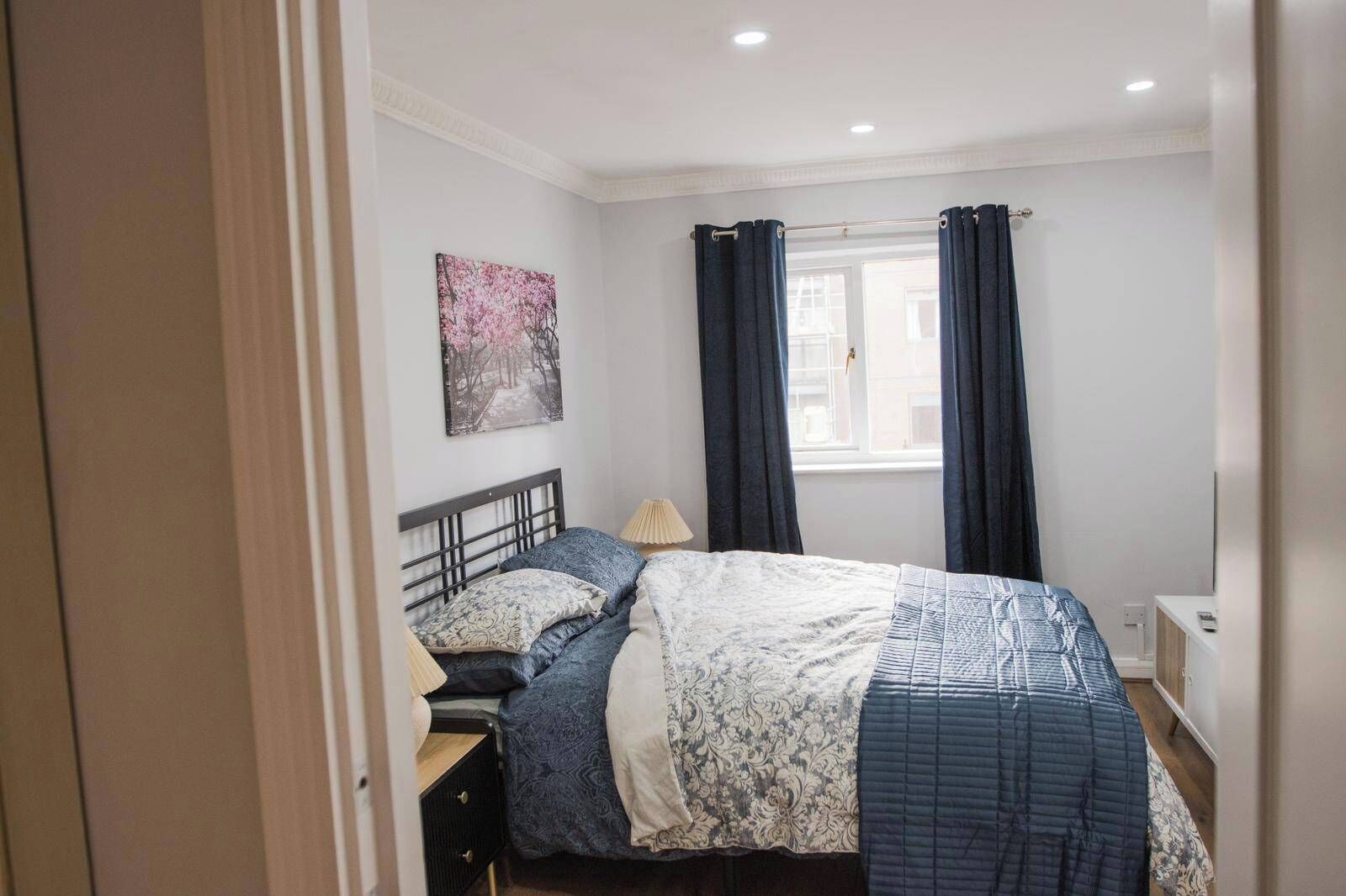 Un dormitorio con cama, mesita de noche, lámpara, ventana con cortinas y un cuadro en la pared.