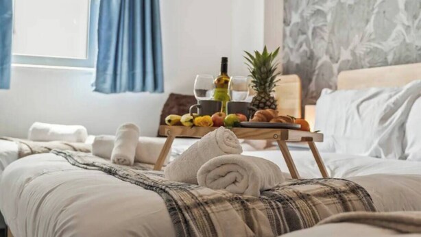 Una cama bien hecha con una manta escocesa, una bandeja con frutas y una botella, y una piña sobre un soporte.