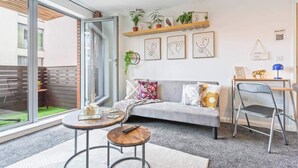 Una sala de estar moderna con un sofá gris, una mesa de centro y un escritorio con una silla. Hay plantas en un estante sobre el sofá y obras de arte en las paredes.