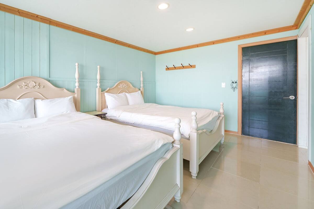 A hotel room with two beds, wooden headboards, a small table, a wall-mounted shelf, and a door.