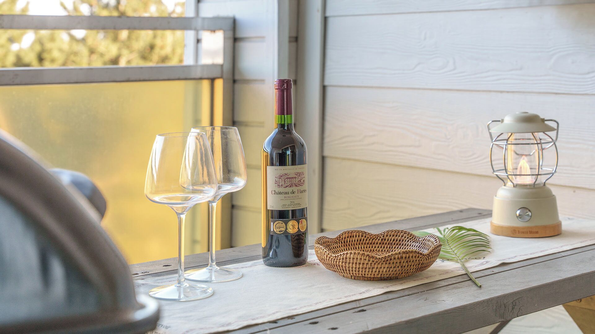 Una botella de vino Chateau de Harlès, dos copas de vino vacías, una cesta tejida y un farol sobre una mesa de madera.