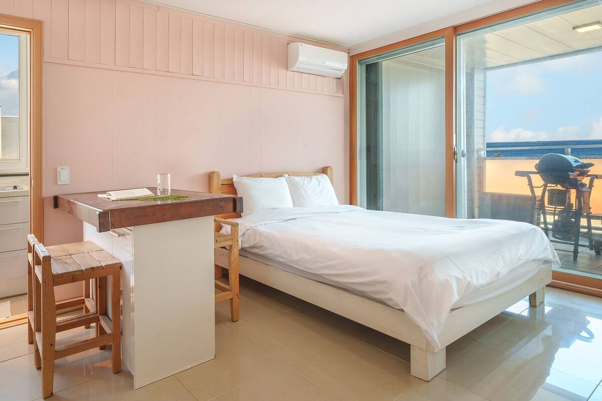 A bedroom with a bed, a small desk, and a chair. There is a window with a view of a building and a balcony with a grill.
