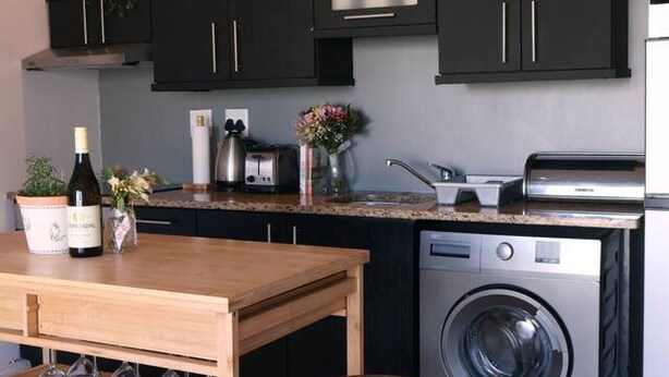 A compact kitchen with dark cabinets, a built-in washing machine, and a wooden table with stools. - Cozy Corner Apartment (Cape Town)
