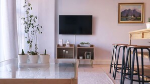 A modern living room with a glass coffee table, potted plants, a flat-screen TV, a painting, and a wooden bar stool area. - Cozy Corner Apartment (Cape Town)