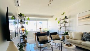 Una sala de estar moderna con un sofá, dos sillones, una mesita de centro y un televisor montado en la pared. Hay estantes con plantas y un ventanal que proporciona luz natural.