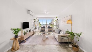 Una sala de estar moderna con un sofá, una mesa de centro y un televisor. Hay plantas y una estantería. La habitación tiene un ventanal con vistas al mar.