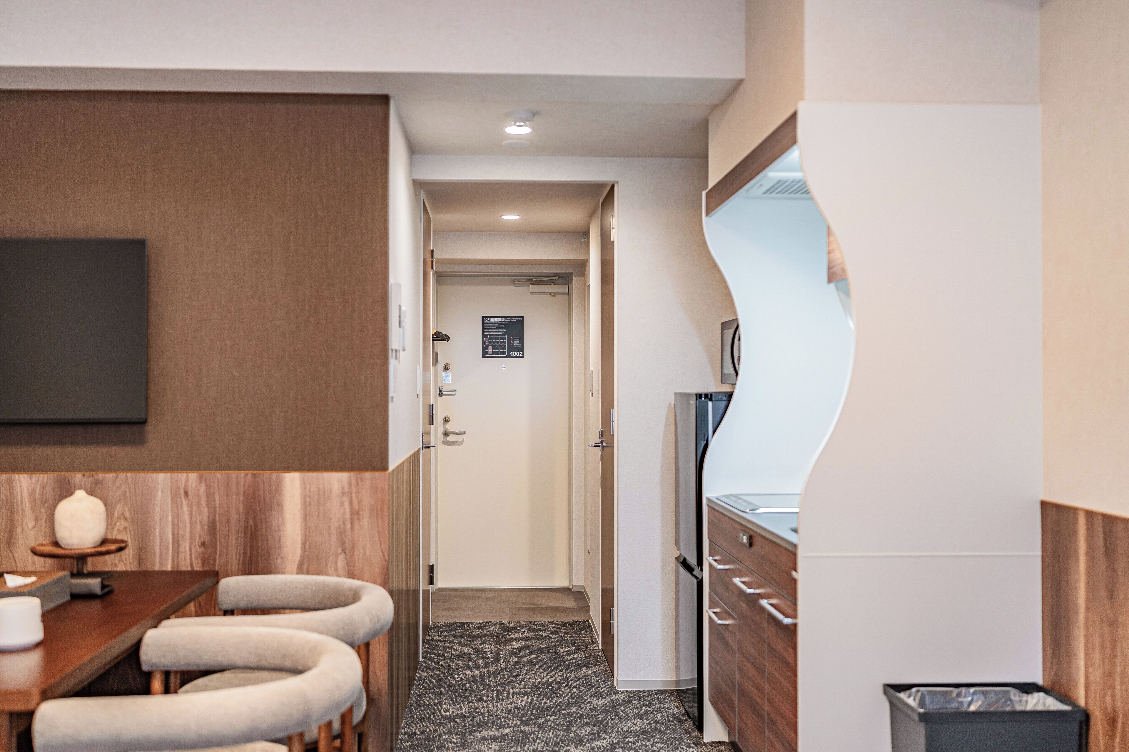 Una habitación de hotel moderna con una zona de bar de madera, un televisor de pantalla plana montado en la pared y una cocina con refrigerador y armarios.