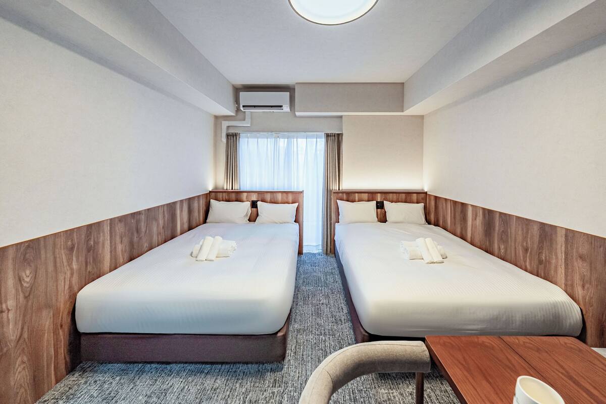 Una habitación de hotel con dos camas, un cabecero de madera, una mesita y una ventana con cortinas.