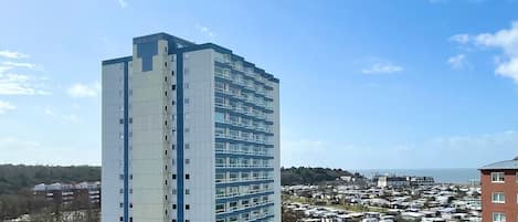A tall, modern hotel with a flat roof and multiple floors. It is surrounded by a parking lot with several cars and a nearby RV park.