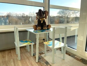 Et værelse med et stort vindue, et hvidt bord med en bamse, og to hvide stole med farverige mønstre.