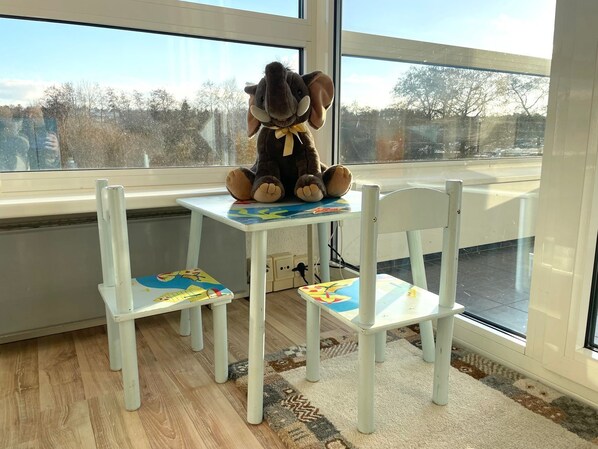 Una habitación con un gran ventanal, una mesa blanca con un peluche, y dos sillas blancas con diseños coloridos.