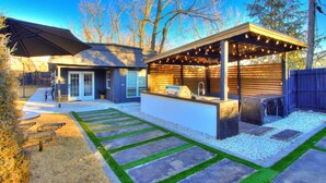 A modern house with a patio area featuring a grill and string lights, surrounded by a well-maintained lawn and a wooden deck.