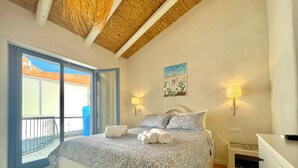 A bedroom with a bed, two bedside lamps, a nightstand, a painting, and a balcony with a view.