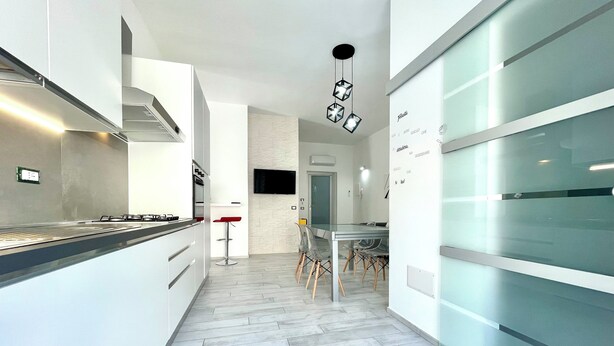 A modern kitchen with a central island, a dining area with a glass table and chairs, and a hallway leading to a living space.