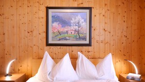 Quarto de hotel com parede revestida de madeira, um quadro emoldurado de uma paisagem, uma cama com roupa branca e duas luminárias de cabeceira.