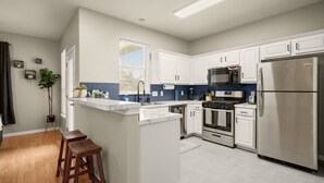 A modern kitchen with white cabinets, stainless steel appliances, and a bar area with stools. - Cozy Retreat With Arcade Haven (Austin)