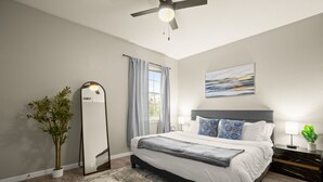 A modern bedroom with a bed, a ceiling fan, a large mirror, a window with curtains, and a painting on the wall. - Cozy Retreat With Arcade Haven (Austin)