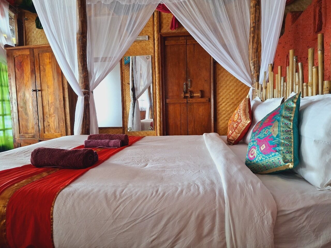 A bed with white linens, a red blanket, and a red towel. There are two pillows with colorful patterns. The room has wooden furniture and a canopy bed.