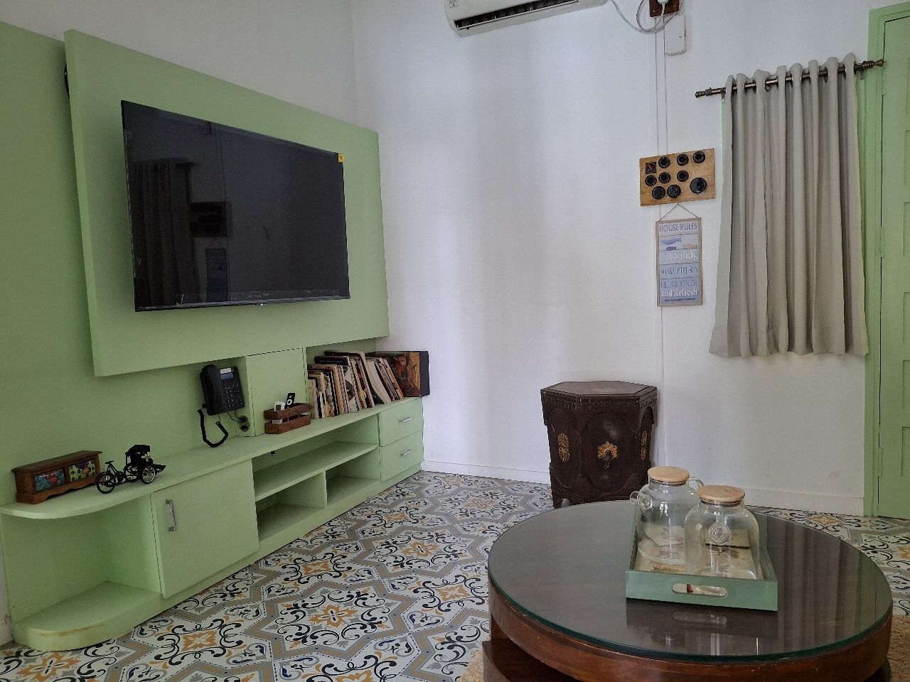 Una sala de estar con un televisor de pantalla plana montado en una pared verde, una mesa redonda de centro con frascos de vidrio, un suelo estampado y un pequeño armario de madera.