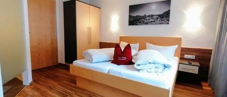A neatly made bed with white bedding and a red pillow, a wooden wardrobe, a nightstand with a lamp, and a framed picture on the wall.