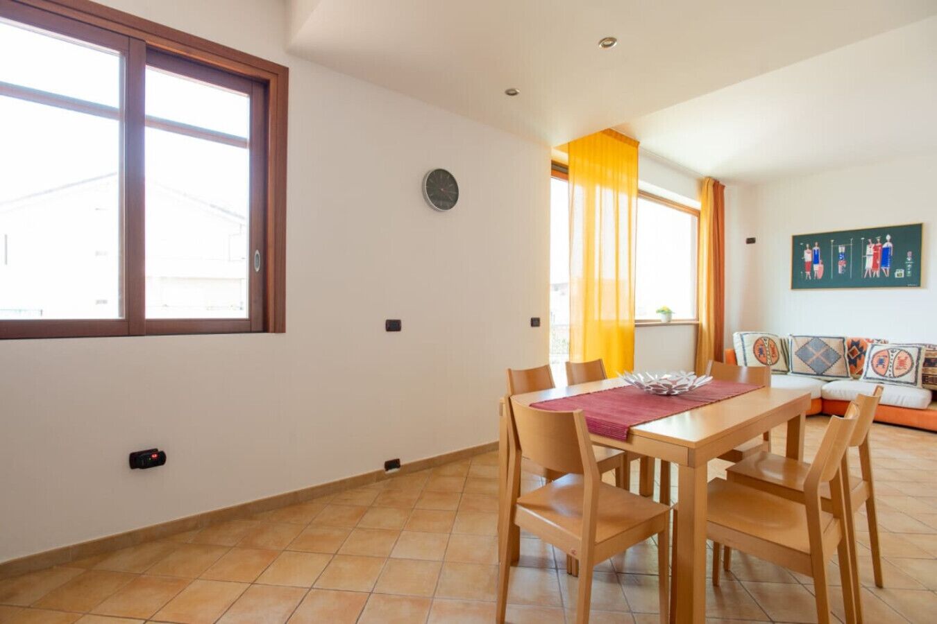 A dining area with a wooden table and chairs, a clock on the wall, and a window with curtains.