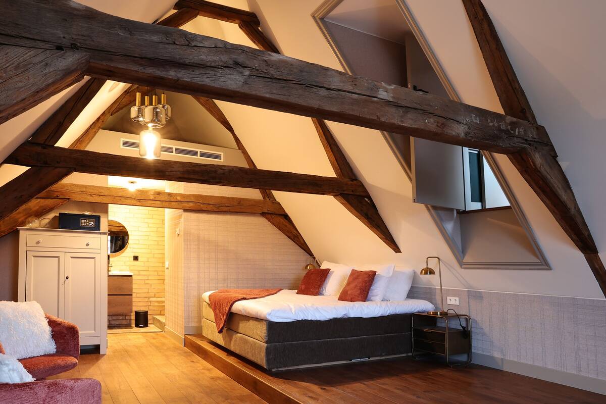 A bedroom with a sloped ceiling, wooden beams, a bed, a nightstand, and a wardrobe.