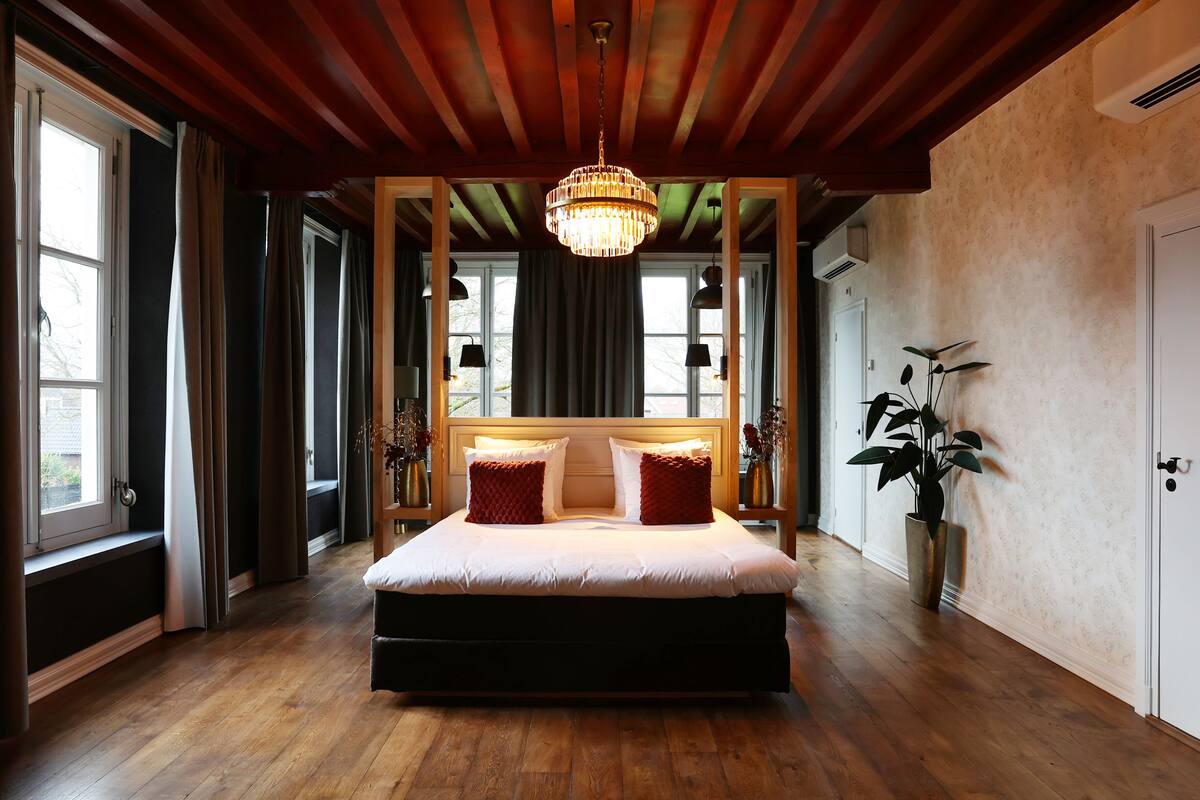 A spacious bedroom with a large bed, wooden ceiling beams, a chandelier, and a potted plant.