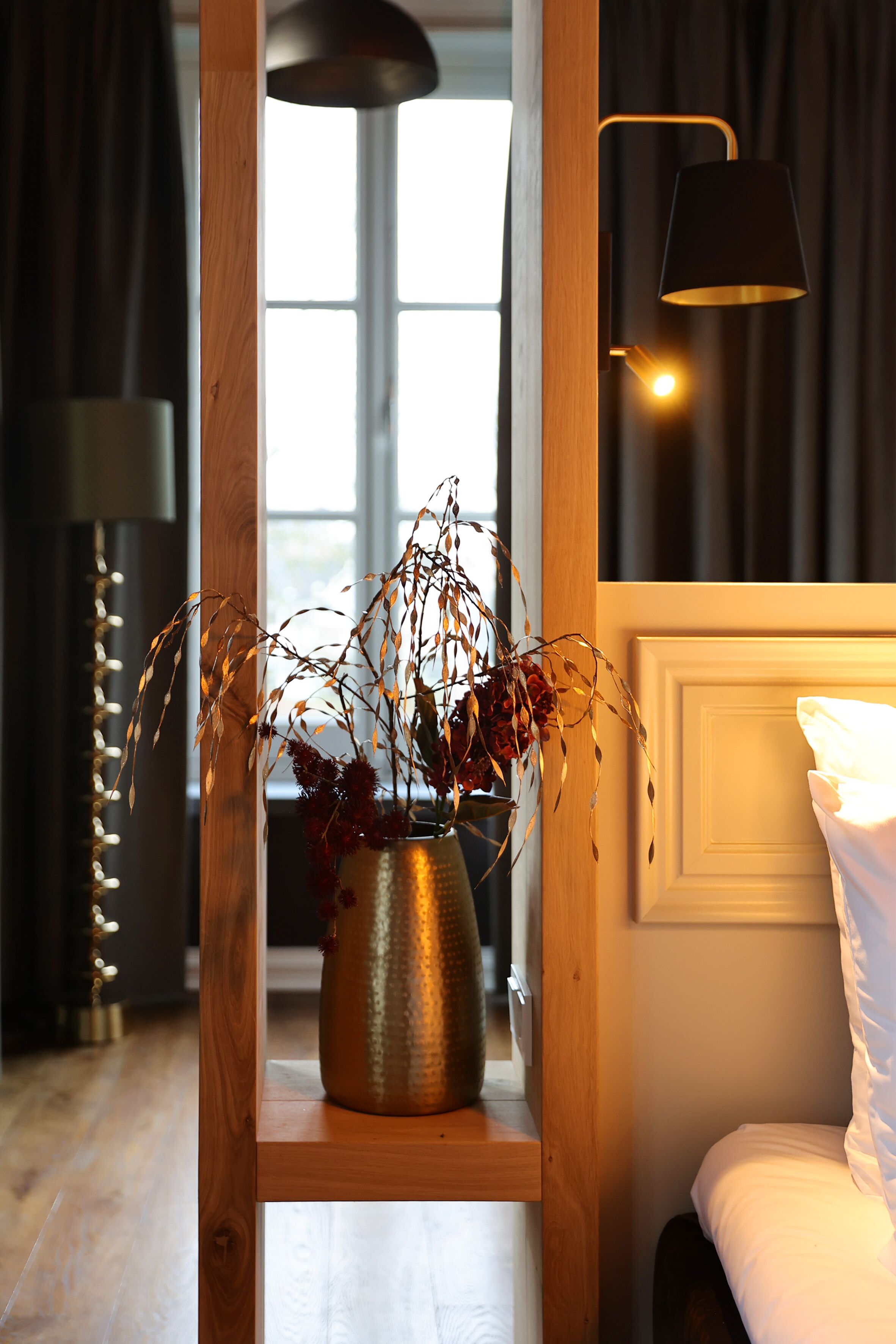 Una estantería de madera con un jarrón de plantas secas, una lámpara de noche y una cama con ropa blanca.