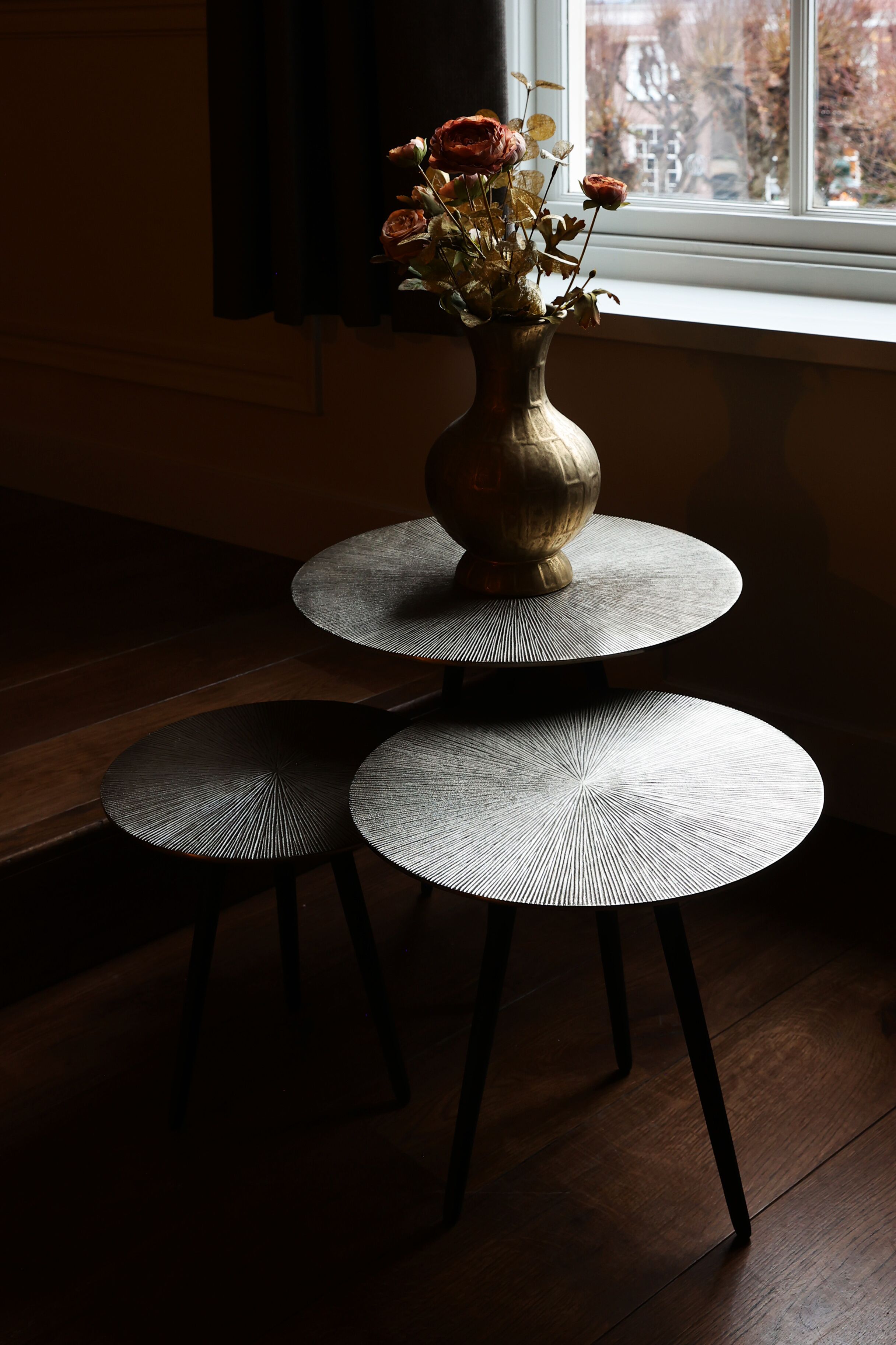 Une table ronde à trois niveaux, sur laquelle est posé un vase de fleurs séchées, est placée près d’une fenêtre donnant sur des arbres et des bâtiments.