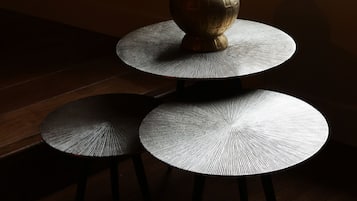 Une table ronde à trois niveaux, sur laquelle est posé un vase de fleurs séchées, est placée près d’une fenêtre donnant sur des arbres et des bâtiments.