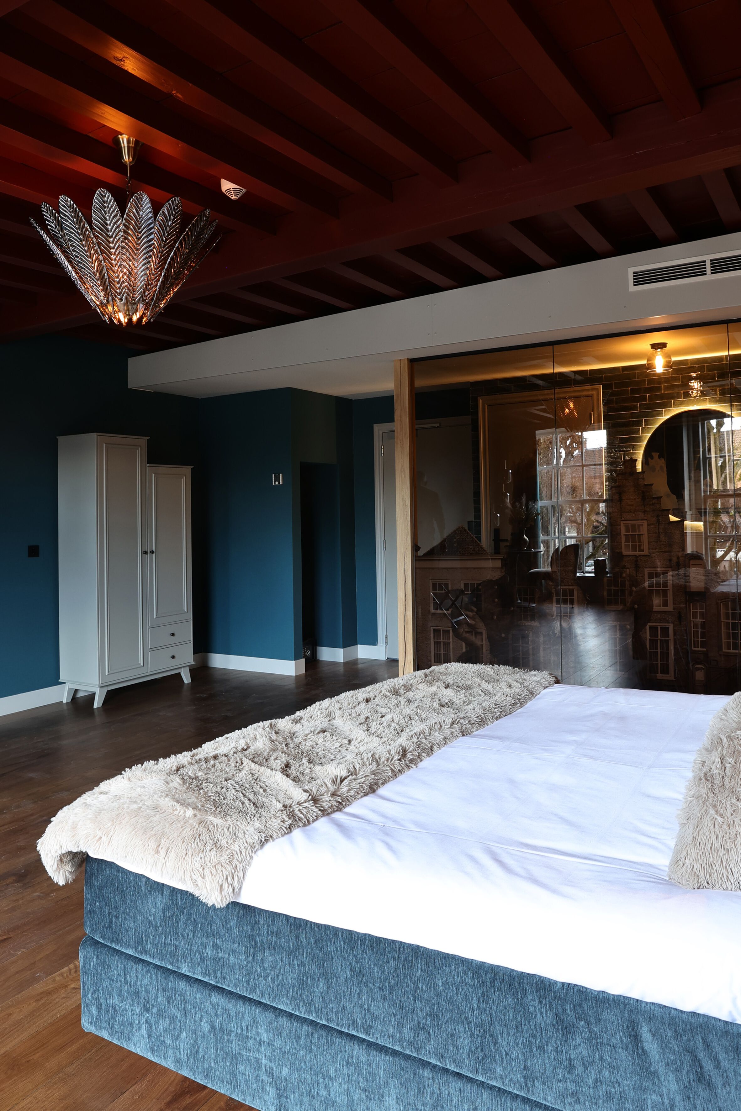 A bedroom with a large bed, wooden beams on the ceiling, a wardrobe, and a view into another room.