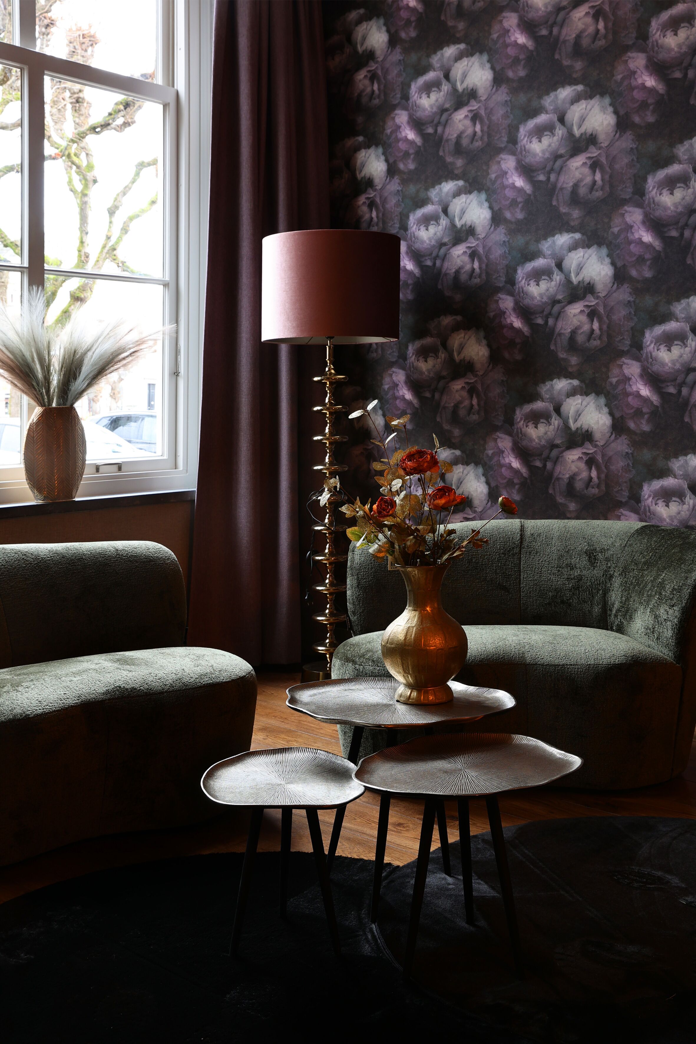 A room with a green sofa, a small round table with a vase of flowers, a tall floor lamp, and a window with curtains.
