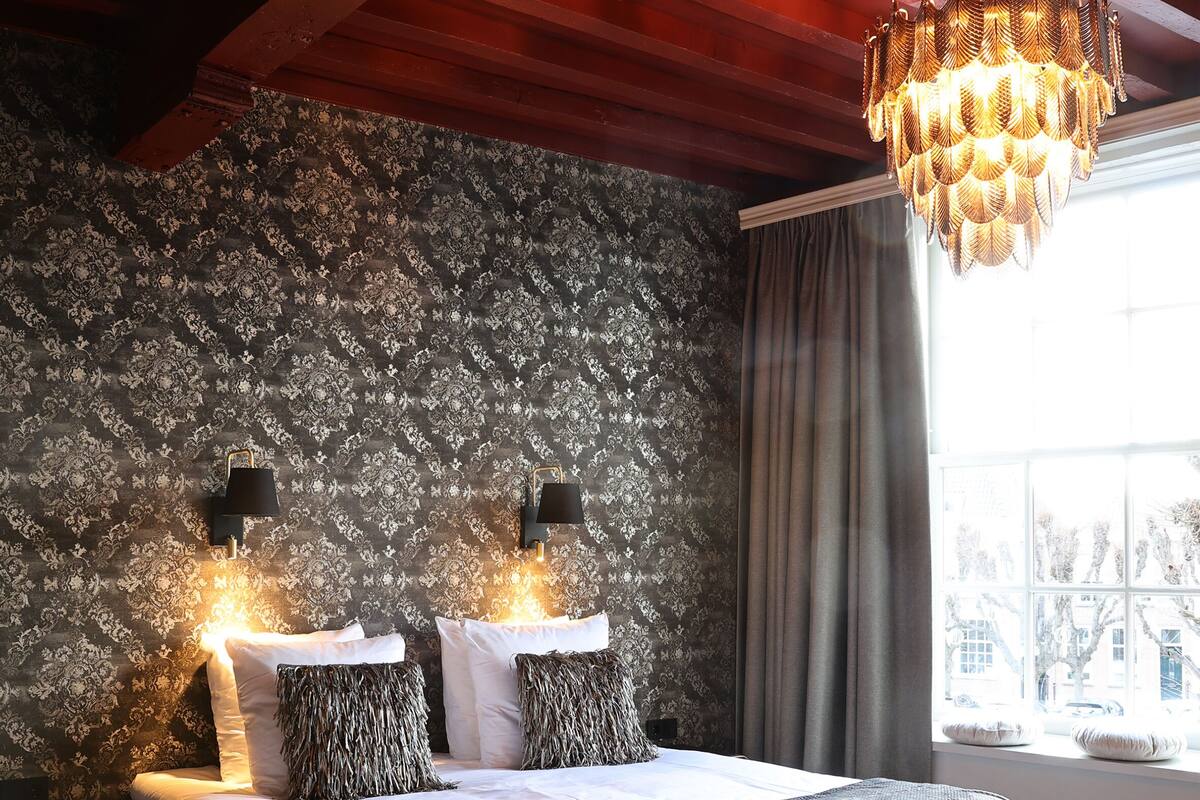 A bedroom with a large bed, wooden beams, a chandelier, and a window with curtains.