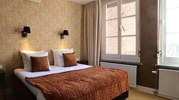 A bedroom with a patterned wallpaper, two bedside lamps, a large window with curtains, and a brown blanket on the bed.