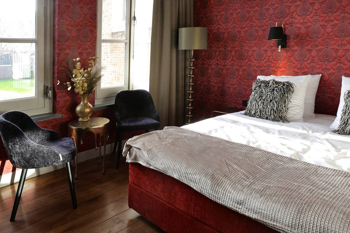 A hotel room with a large bed, two chairs, a small table, a vase with flowers, and a lamp.