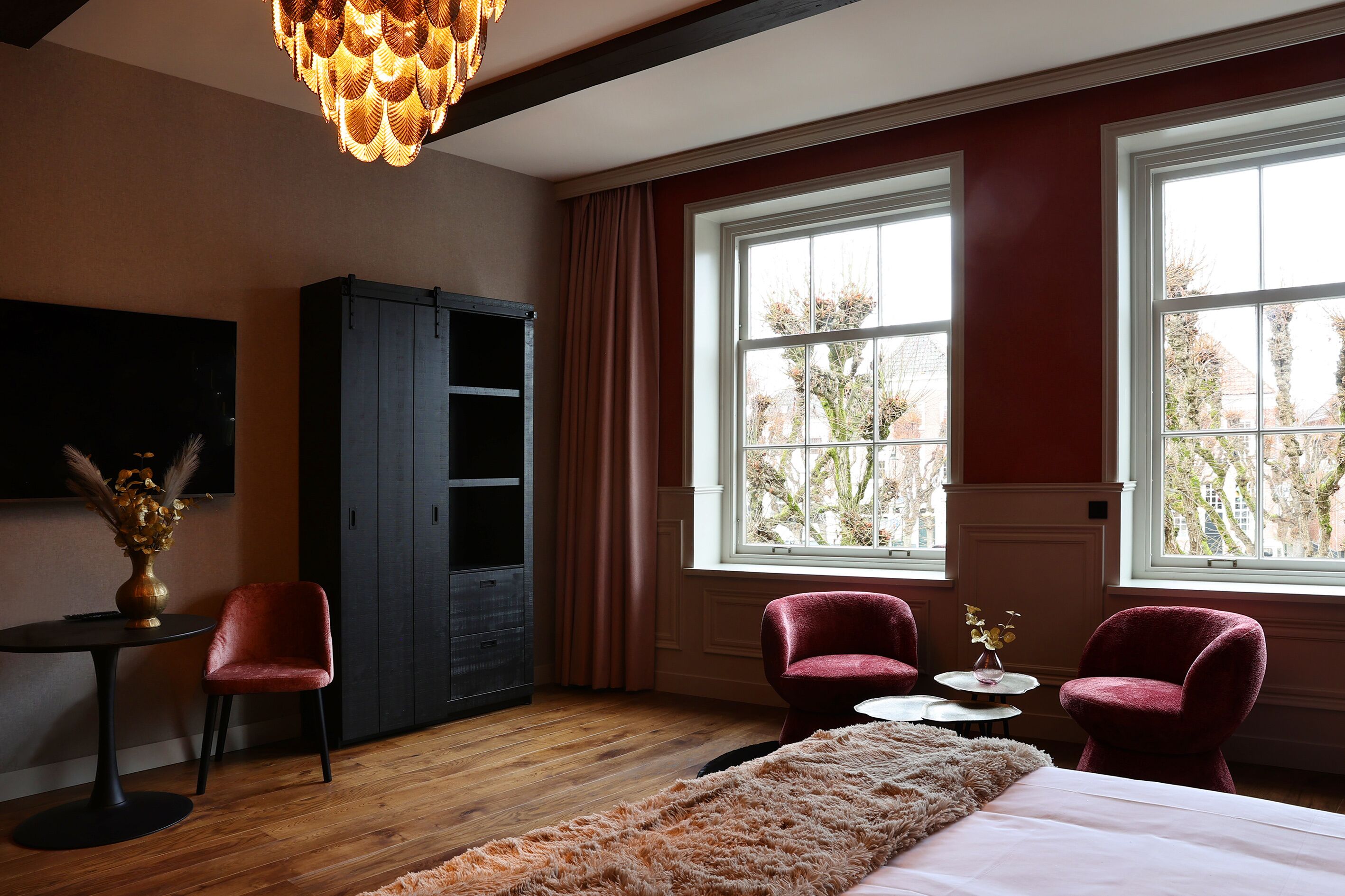 A bedroom with a large bed, two red armchairs, a small table with a vase, a dark wardrobe, and a large window with a view of trees.