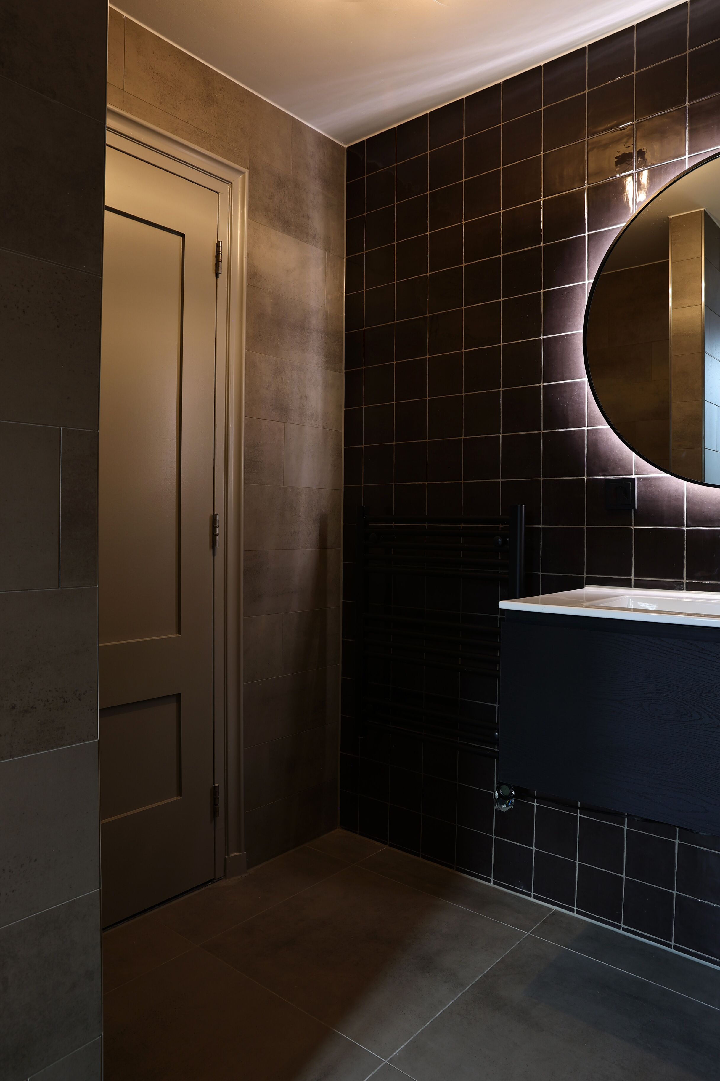 A bathroom with a dark tiled wall, a round mirror, and a white door.