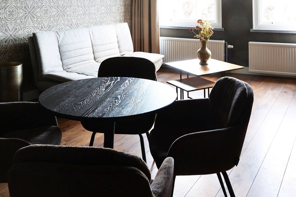 A modern living room with a high ceiling, exposed beams, a patterned wallpaper, a sofa, a round table, and two chairs.