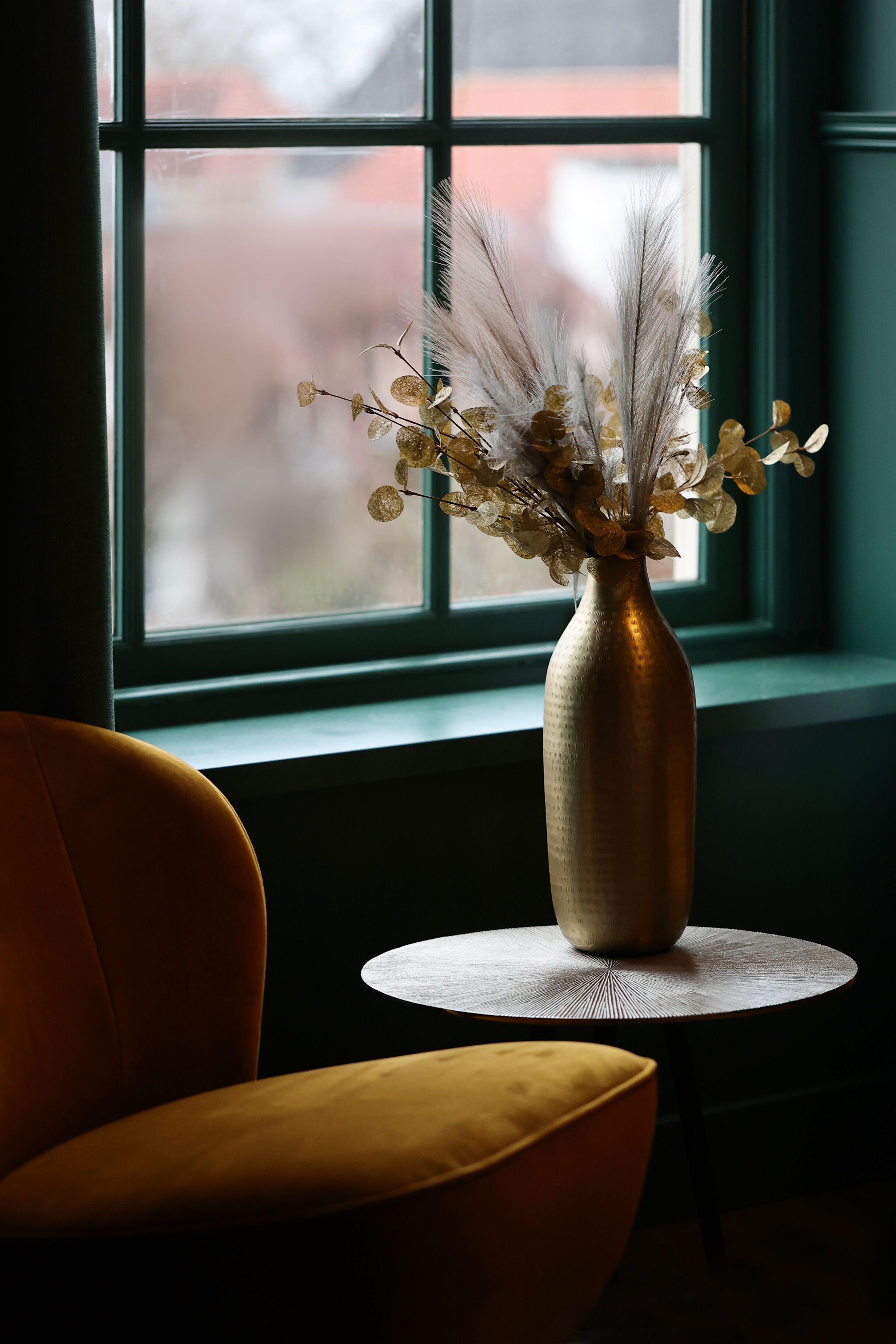 Een kamer met een raam, een vaas met gedroogde bloemen, een ronde tafel en een gele stoel.