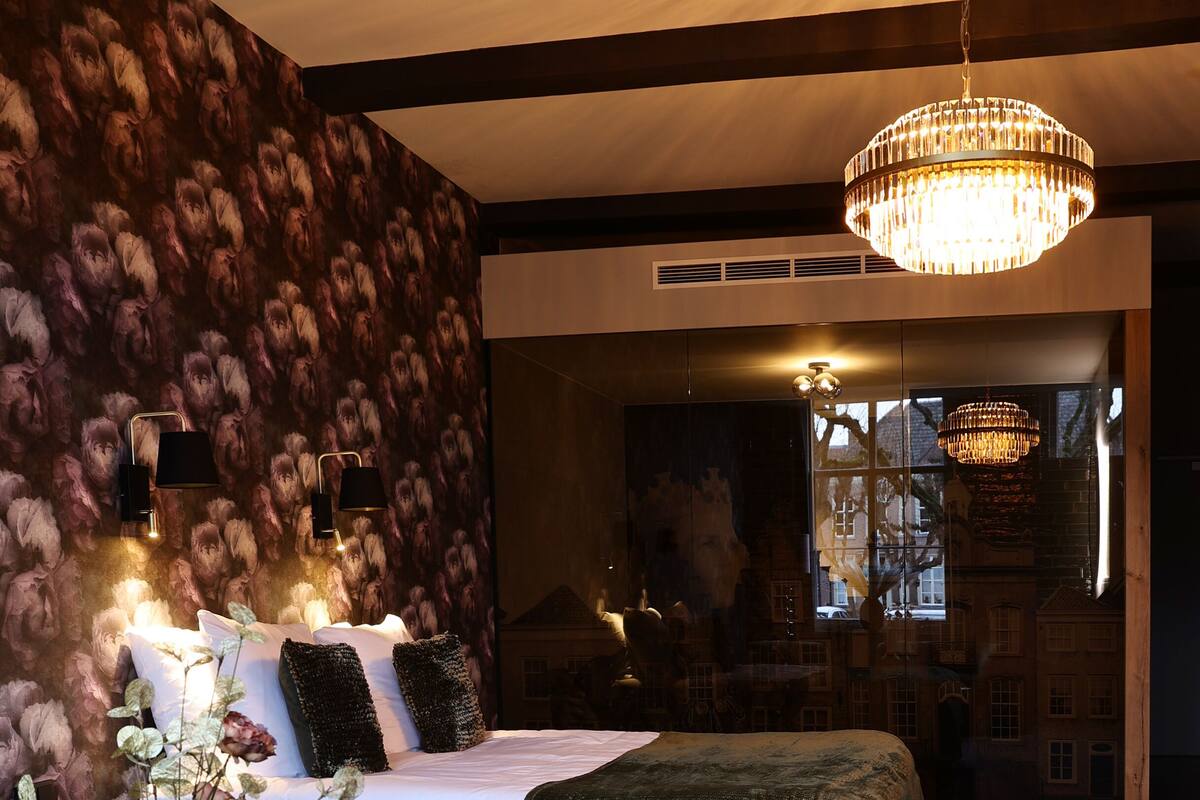 A bedroom with a large bed, a chandelier, a vase with flowers, and a view of a cityscape through a window.