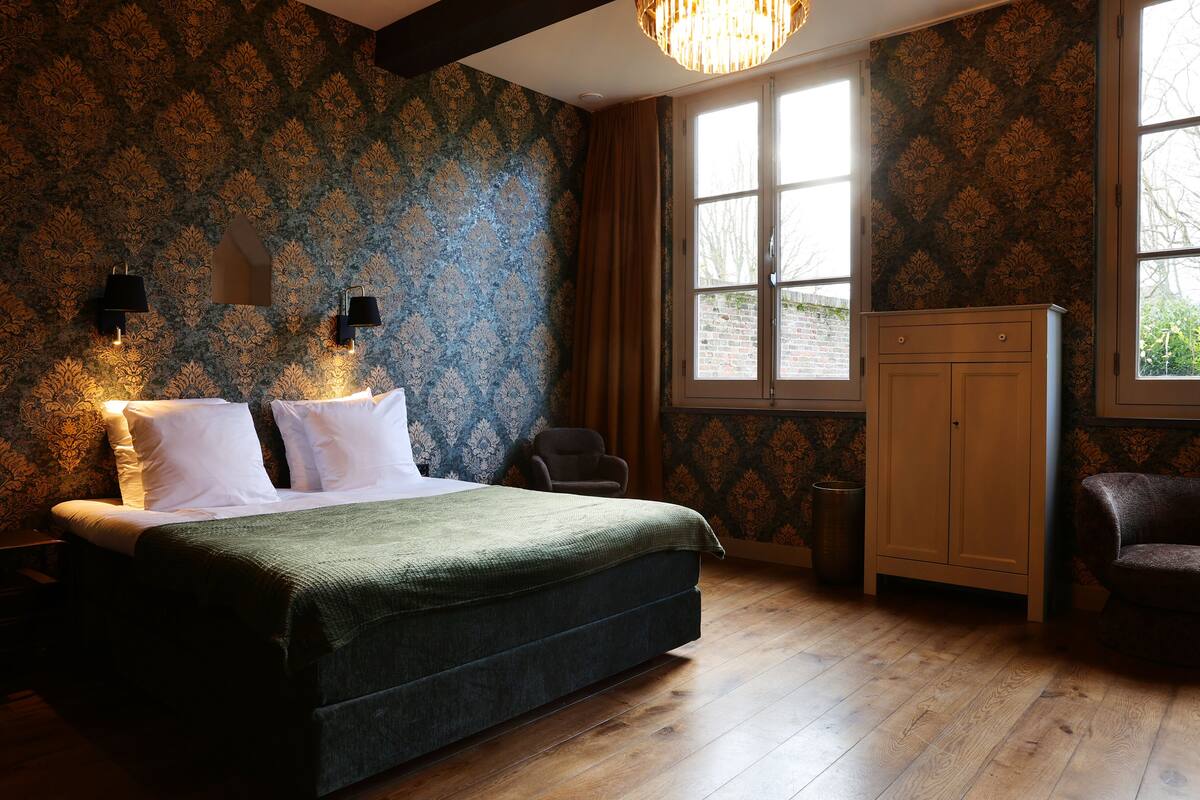 A bedroom with a large bed, wooden flooring, patterned wallpaper, a wardrobe, and a window with curtains.