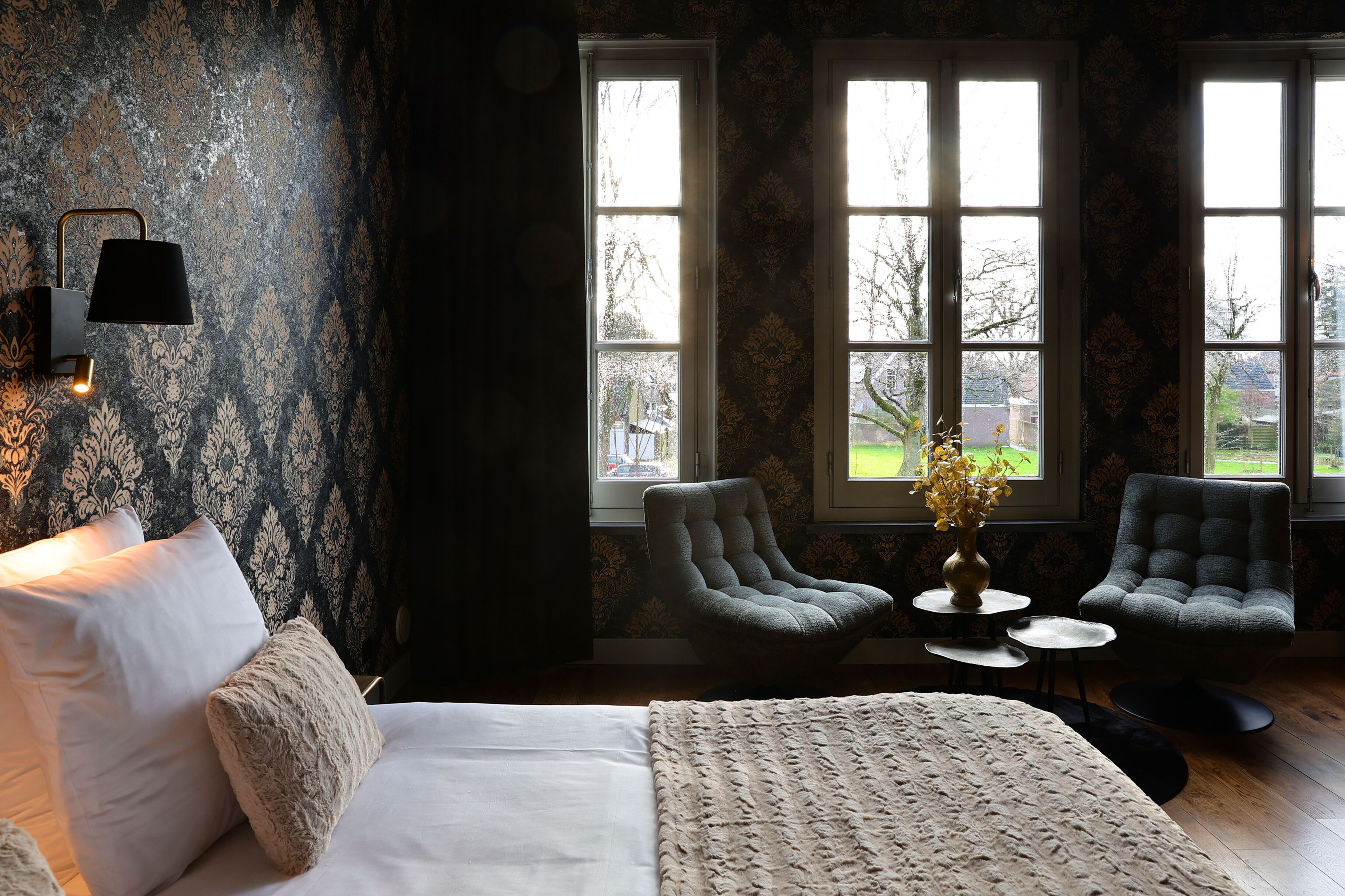 A bedroom with a bed, two armchairs, a small table, and a lamp. There are two windows with a view of the outside.