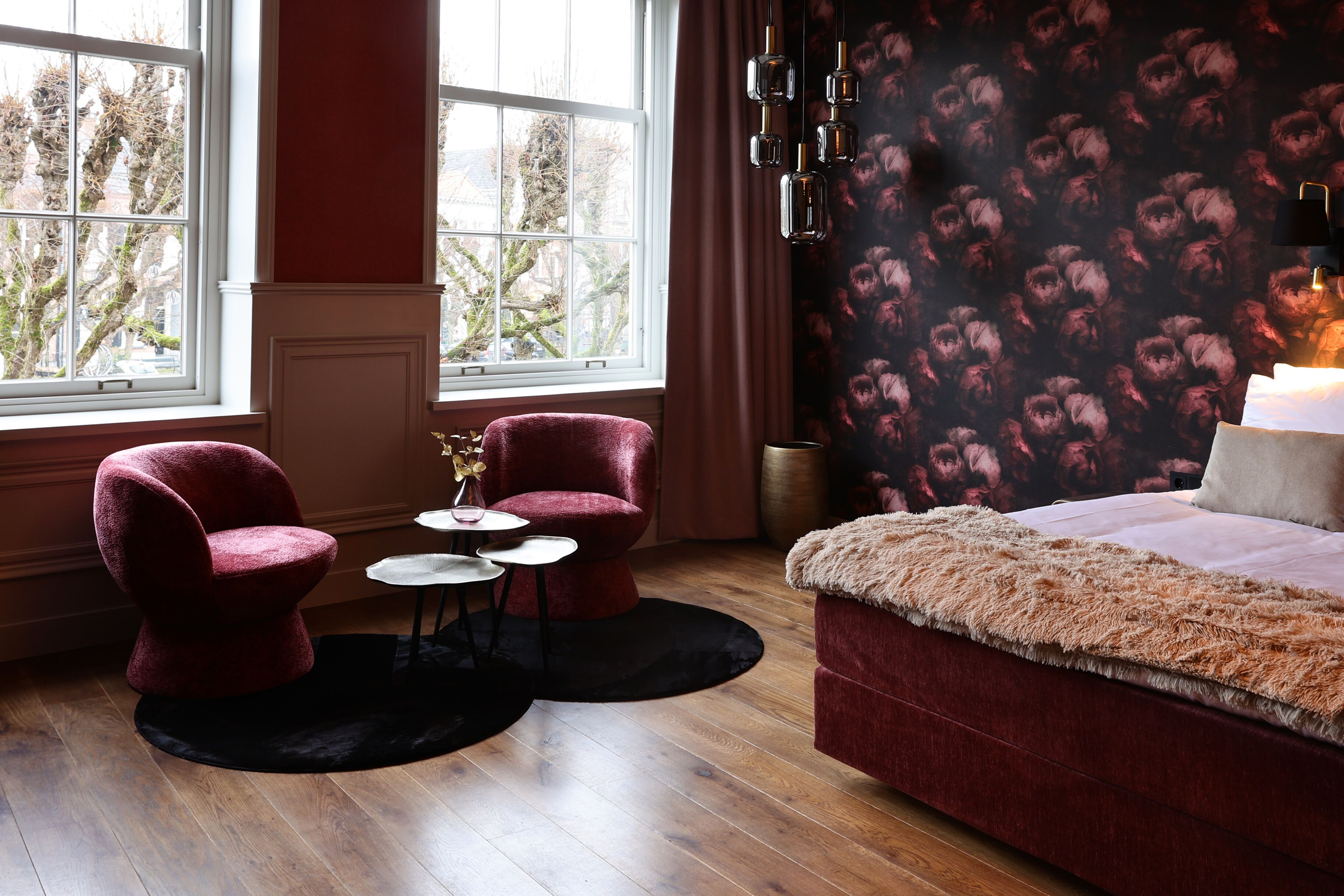 Een slaapkamer met een groot raam, twee rode fauteuils, een kleine ronde tafel en een bed met een zachte deken.
