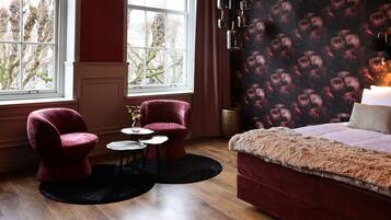 Een slaapkamer met een groot raam, twee rode fauteuils, een kleine ronde tafel en een bed met een zachte deken.