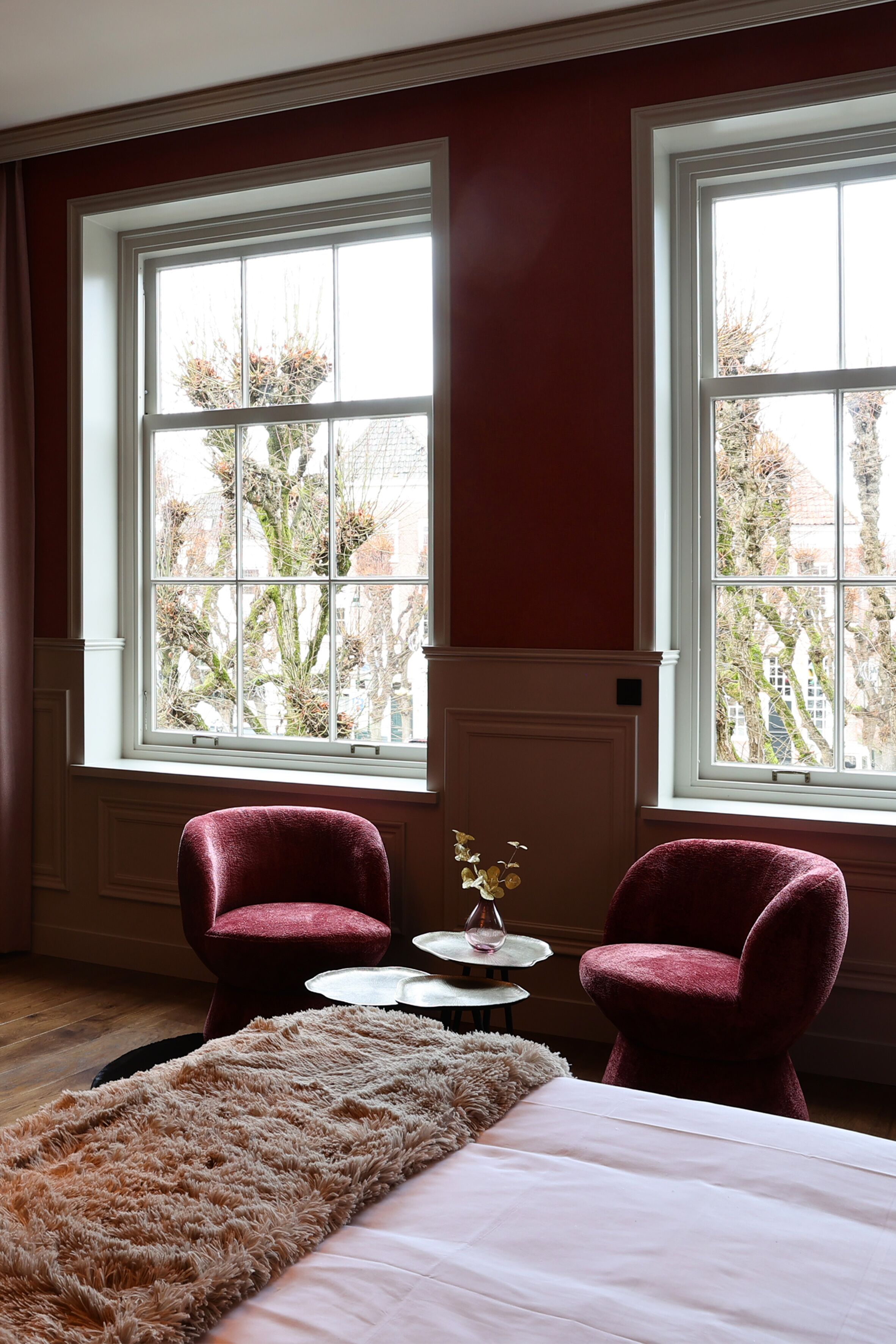 A room with two large windows, a bed, a fluffy rug, and two chairs.