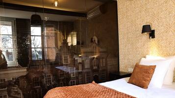 Habitación de hotel con una cama grande, una manta marrón, un almohadón blanco y una mesita de noche con una lámpara.