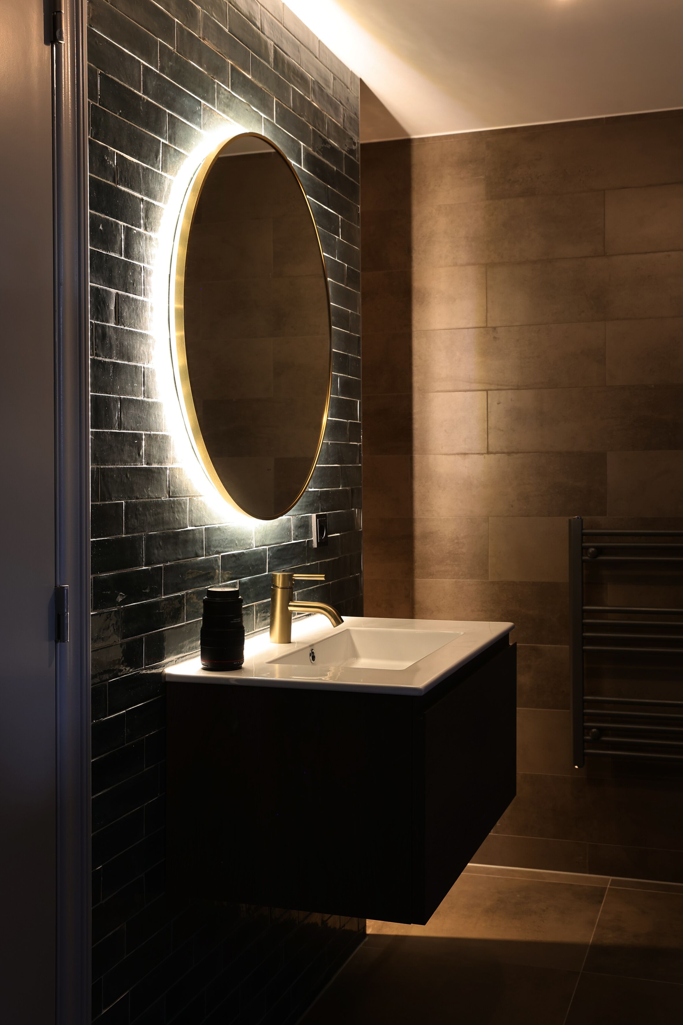 Un baño moderno con pared de azulejos negros, un espejo con marco dorado, un lavabo blanco y un perchero para toallas.