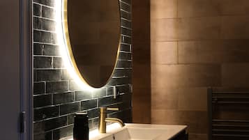 Un baño moderno con pared de azulejos negros, un espejo con marco dorado, un lavabo blanco y un perchero para toallas.