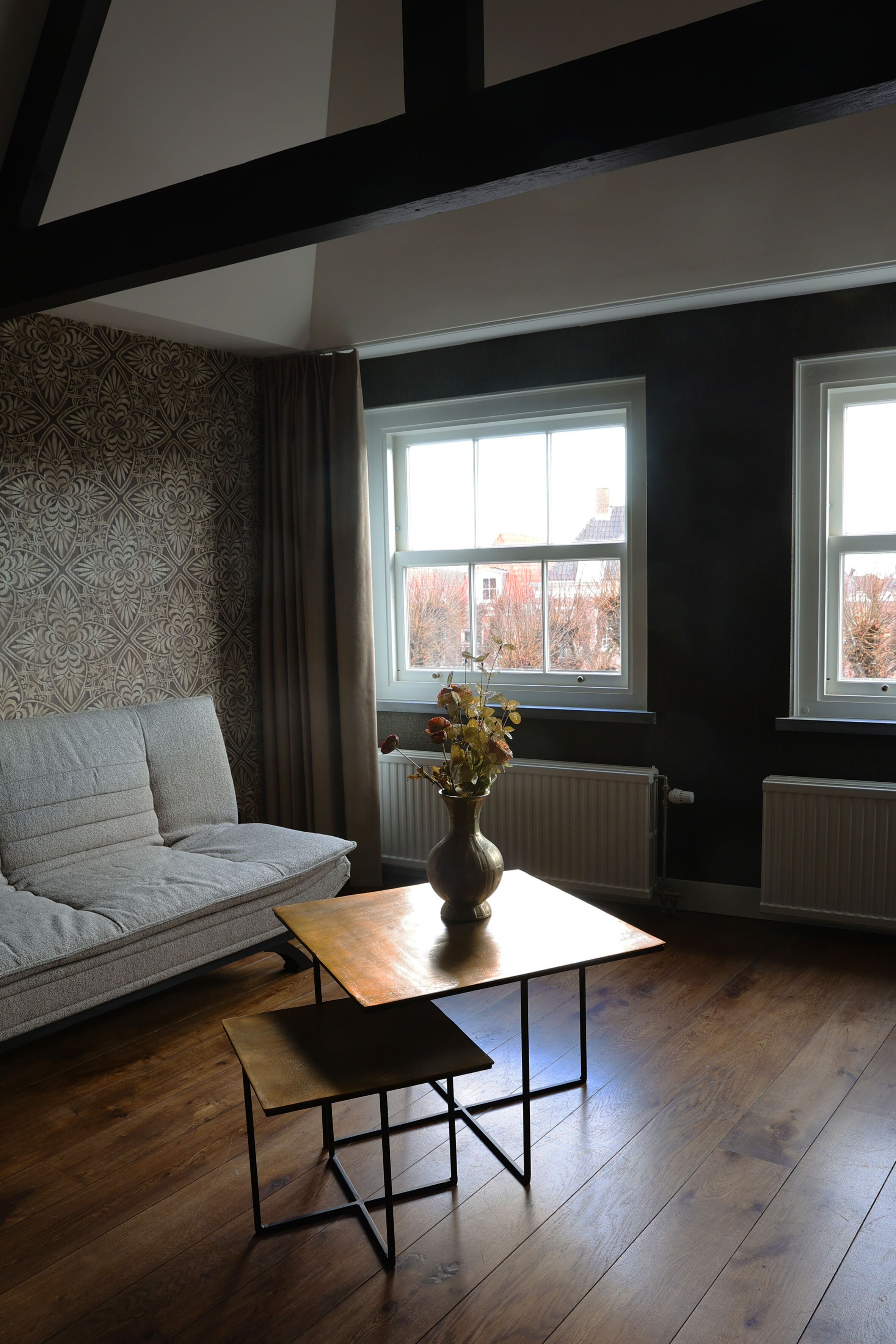 A room with a sofa, a wooden table, and a vase with flowers. There are two windows with curtains and a radiator.