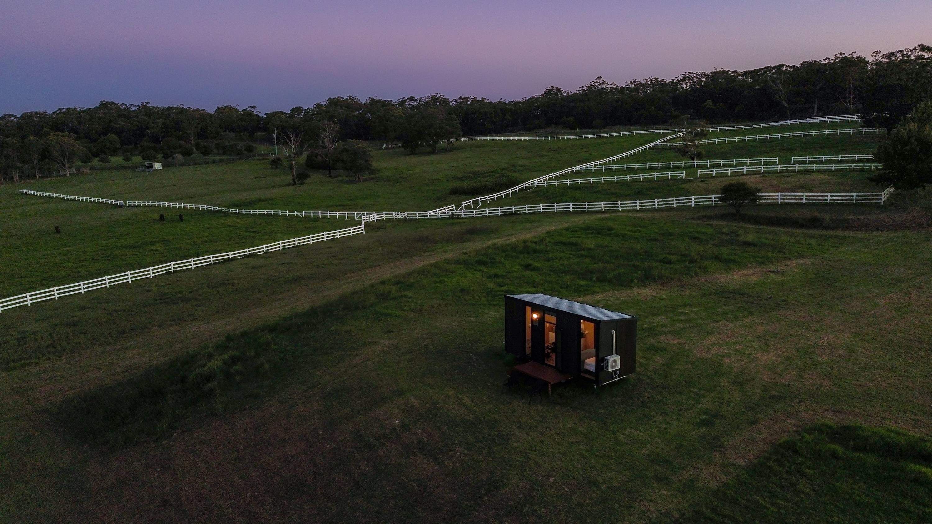 Une cabane isolée et rustique, composée d’une seule pièce, entourée de clôtures blanches et située dans un paysage vallonné.